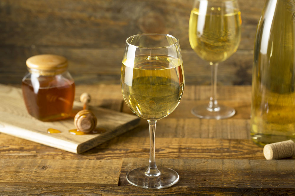 Zwei Gläser mit goldfarbenem Met neben Honigglas und Flasche auf einem Holztisch
